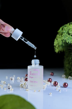 A hand holds a glass dropper above a frosted bottle labeled Jasmin Botanical Glow, with a single clear drop hanging between the dropper and bottle. The scene is styled on a reflective white surface with scattered pearl-like beads, a dark black background, and green foliage props.