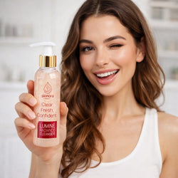 A smiling woman with wavy brown hair holds up a bottle of Skimora Clear Fresh Confident Lumine Radiance Cleanser. The bottle is a frosted peachy-pink color with a gold pump collar and a white pump dispenser.